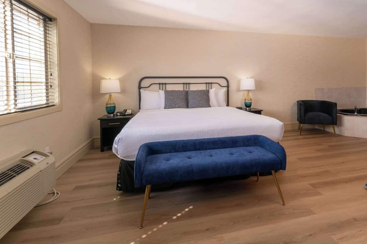 A modern bedroom with a large bed, bedside lamps, a blue bench, a small desk, and a view of the outdoors through the window.