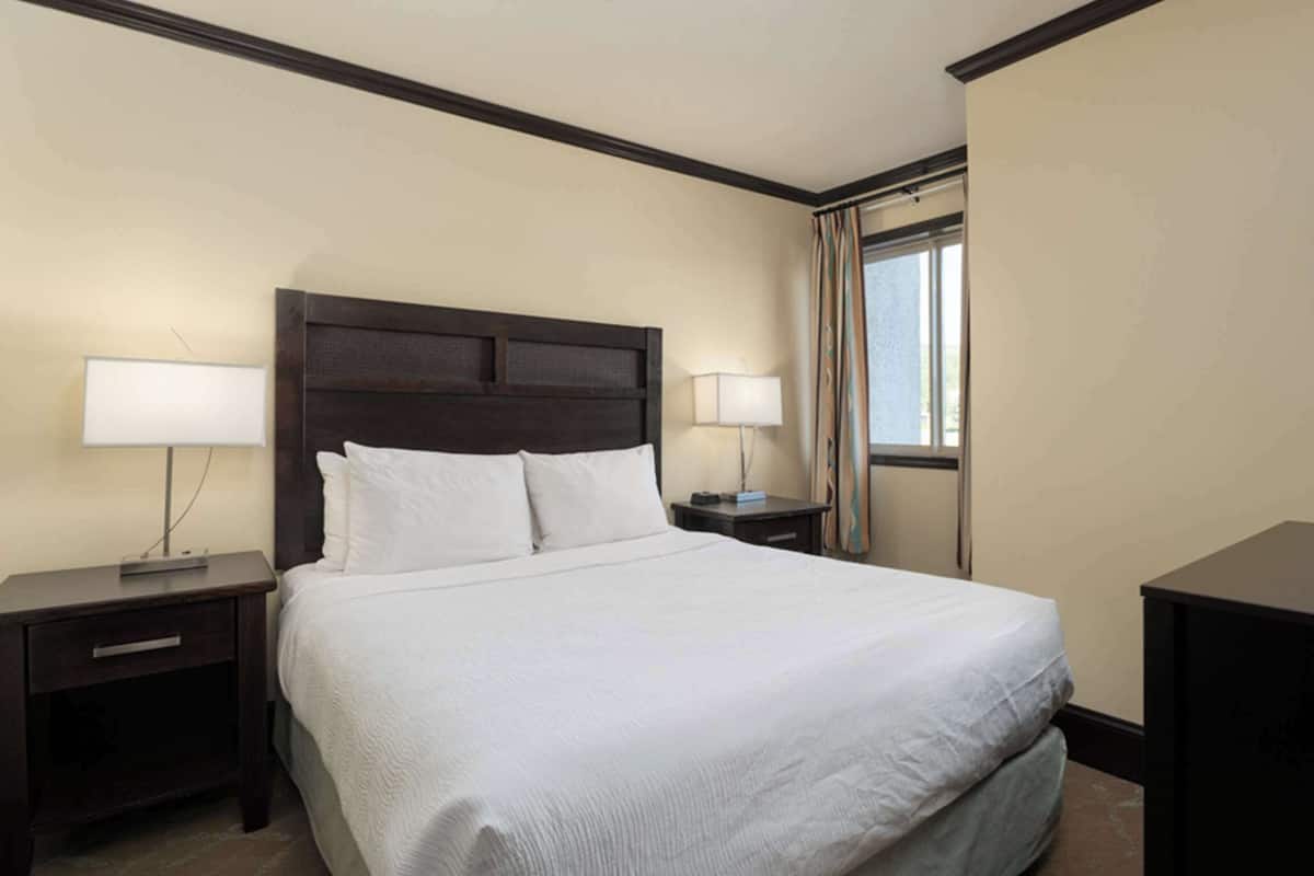 A hotel room with a large bed, two bedside tables with lamps, a dark headboard, and a window with curtains.