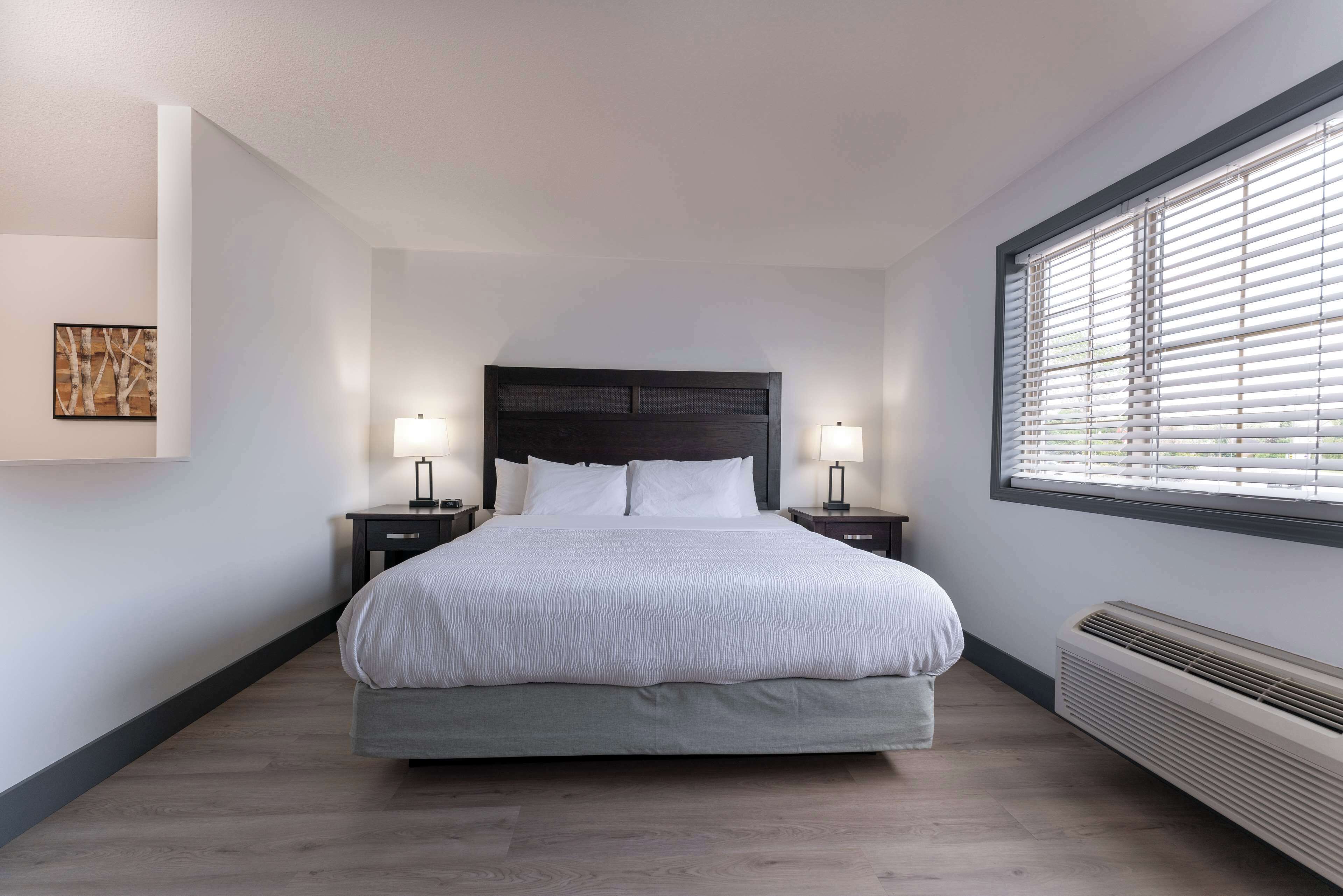 Een moderne slaapkamer met een groot bed, twee nachtkastjes met lampen, een kunstwerk aan de muur en een raam met zonwering.