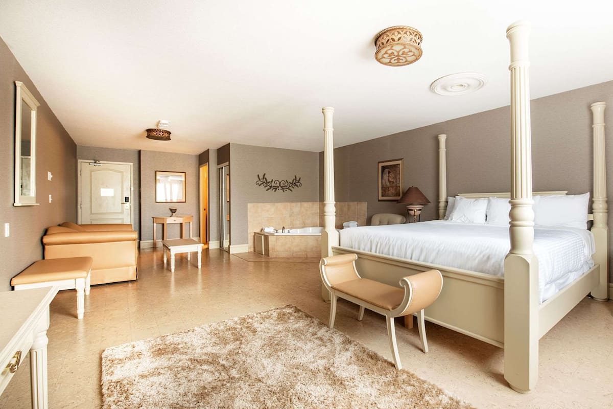 A spacious bedroom with a four-poster bed, a sitting area with a bench and chairs, a small table, and a nightstand with a lamp.