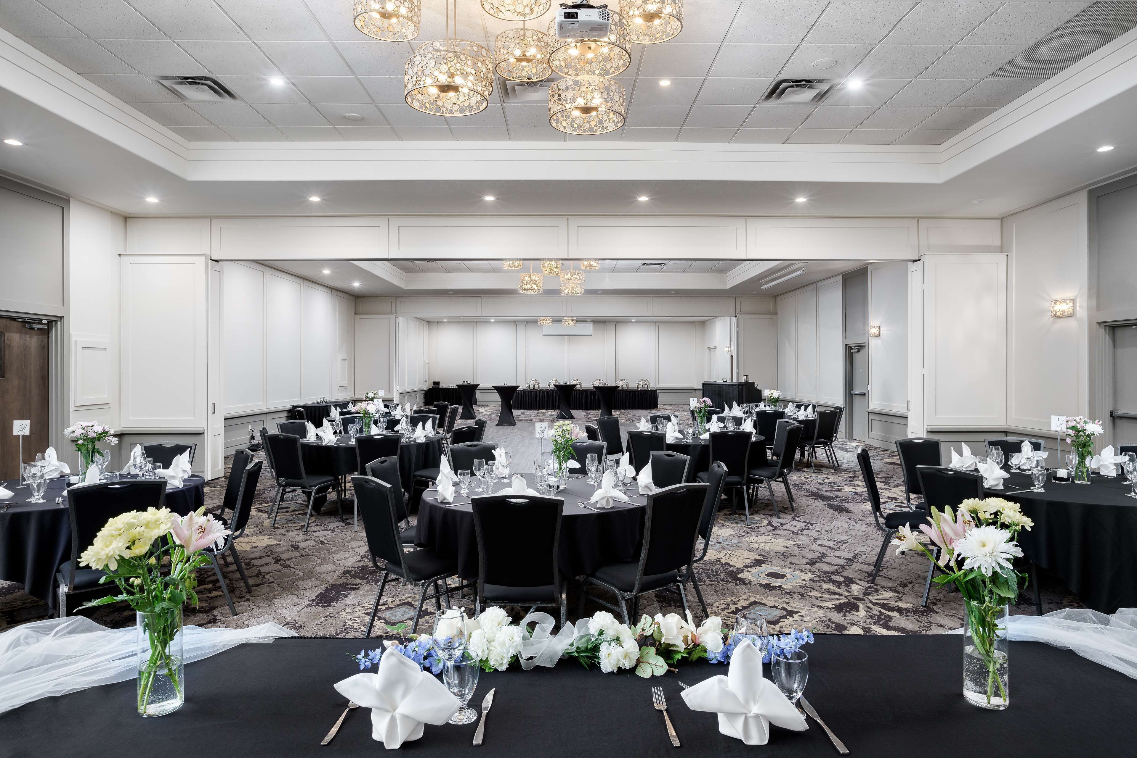 Un salón de banquetes con mesas redondas preparadas para eventos, con manteles negros, servilletas blancas y centros de mesa florales.