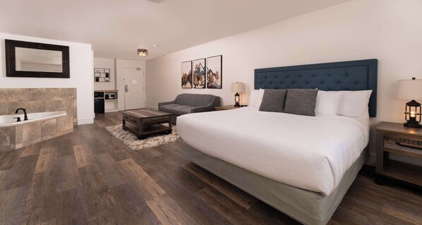 A hotel room with a large bed, a sitting area with a gray sofa, a small table, a wall-mounted mirror, and a bathtub.