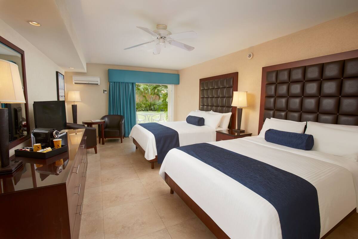 A hotel room with two beds, a desk with a computer, a television, and a balcony with a view of greenery.