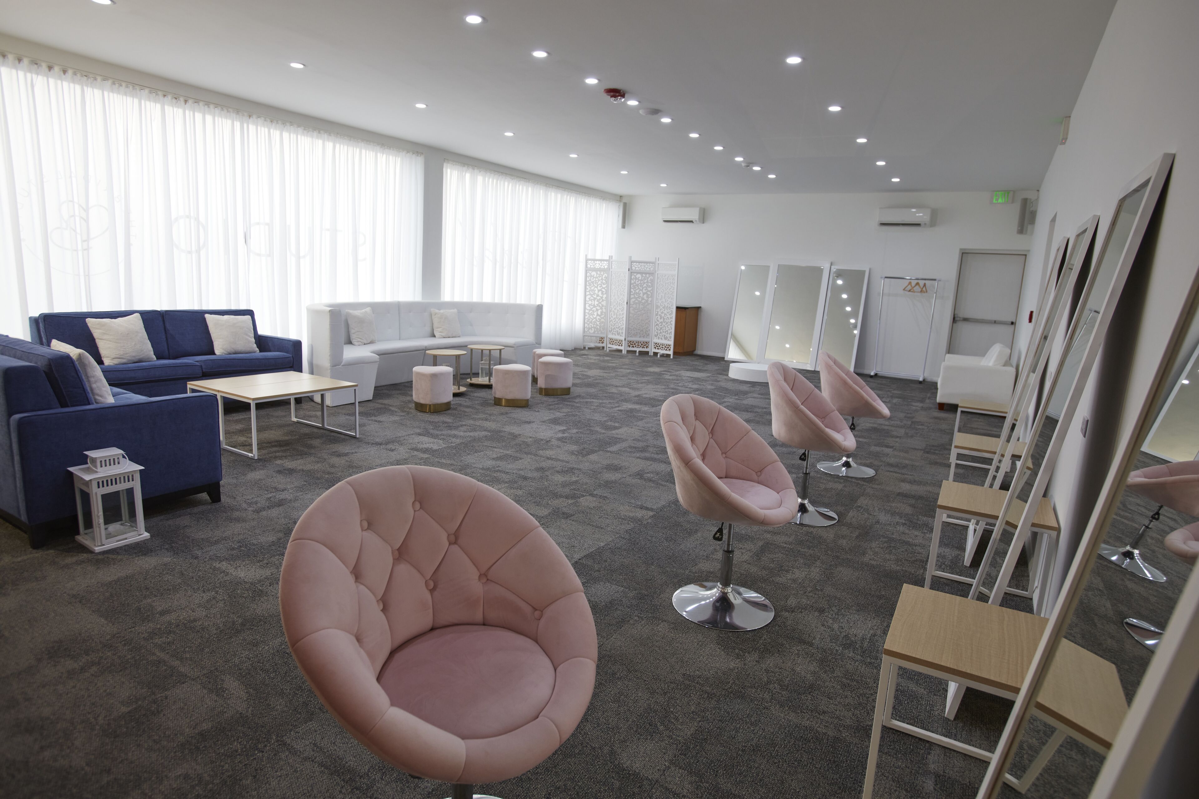 A modern hotel room with a blue sofa, a white sofa, a pink chair, and a wooden chair. There are multiple mirrors and a white partition.