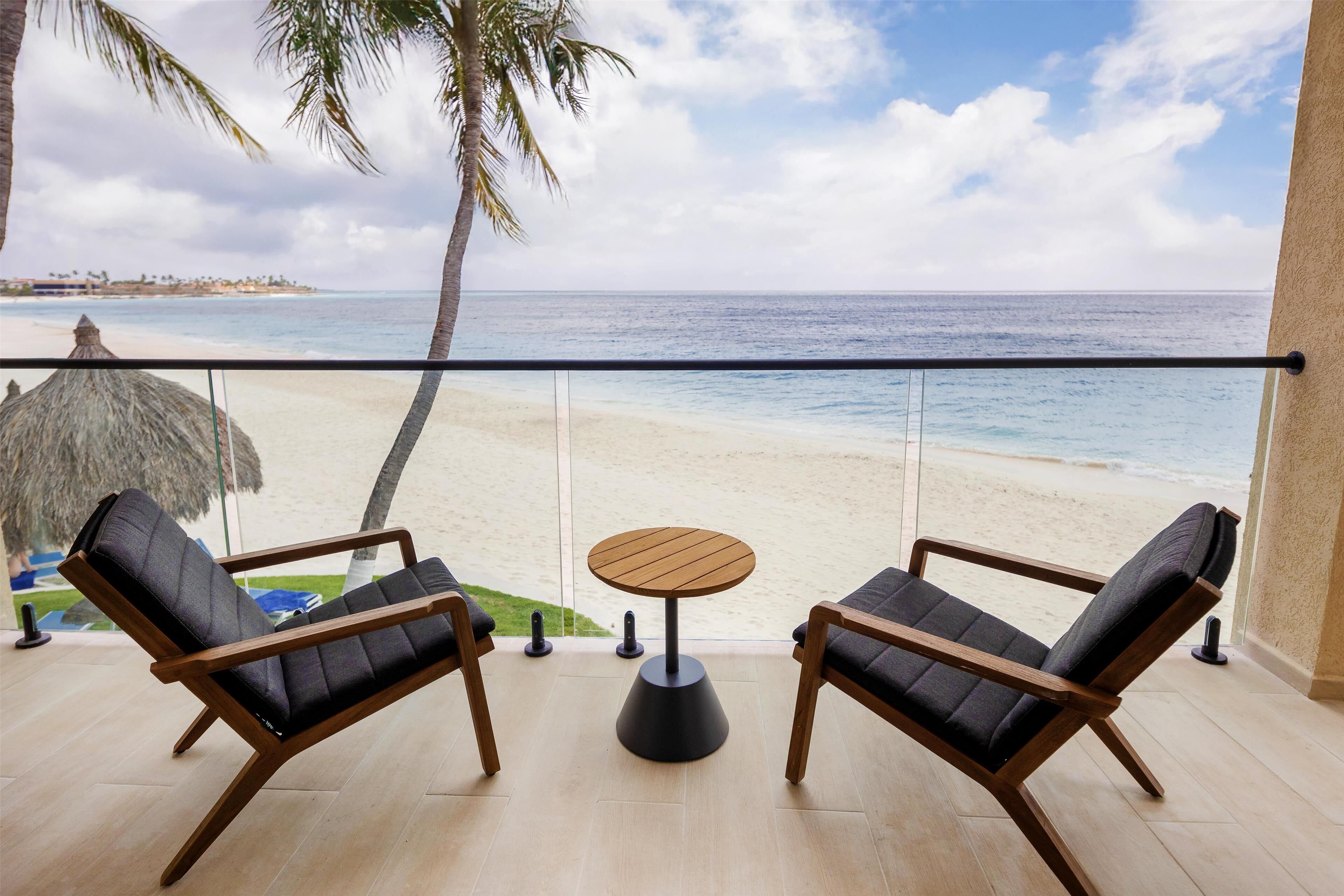 Un balcón con dos sillones de descanso y una pequeña mesa redonda, con vista a una playa donde se ve una palmera y una cabaña de techo de paja al fondo.