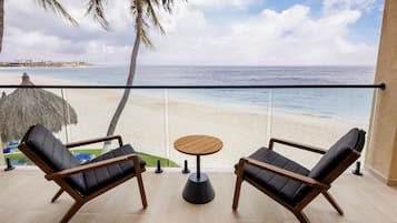 Un balcón con dos sillones de descanso y una pequeña mesa redonda, con vista a una playa donde se ve una palmera y una cabaña de techo de paja al fondo.