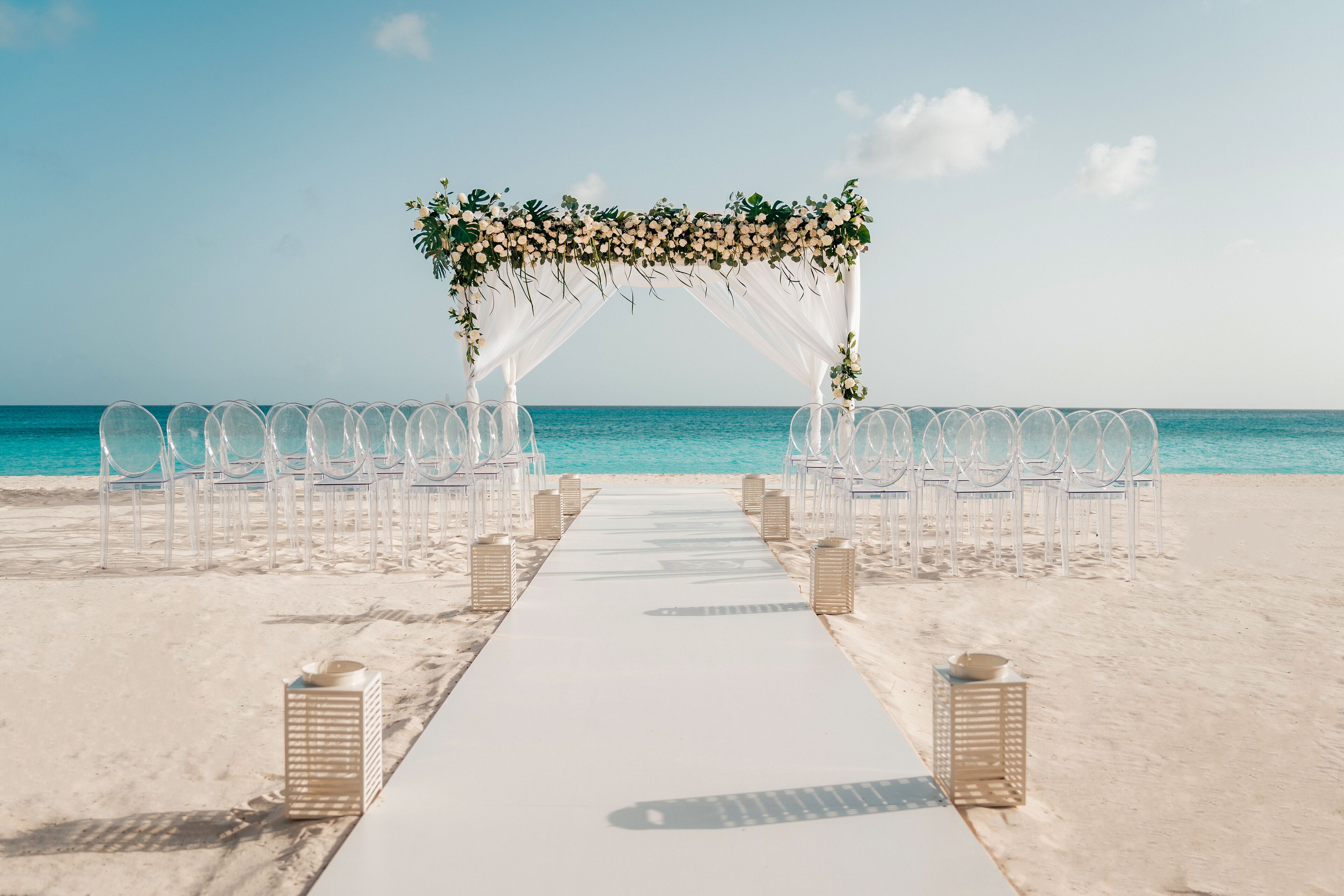 A beach wedding setup with a white aisle, chairs, and a floral arch.