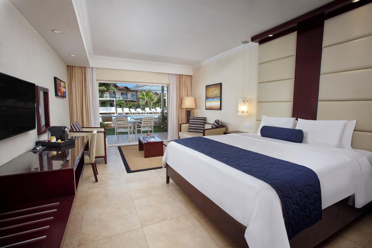 A hotel room with a large bed, a desk, a TV, and a balcony with outdoor seating.
