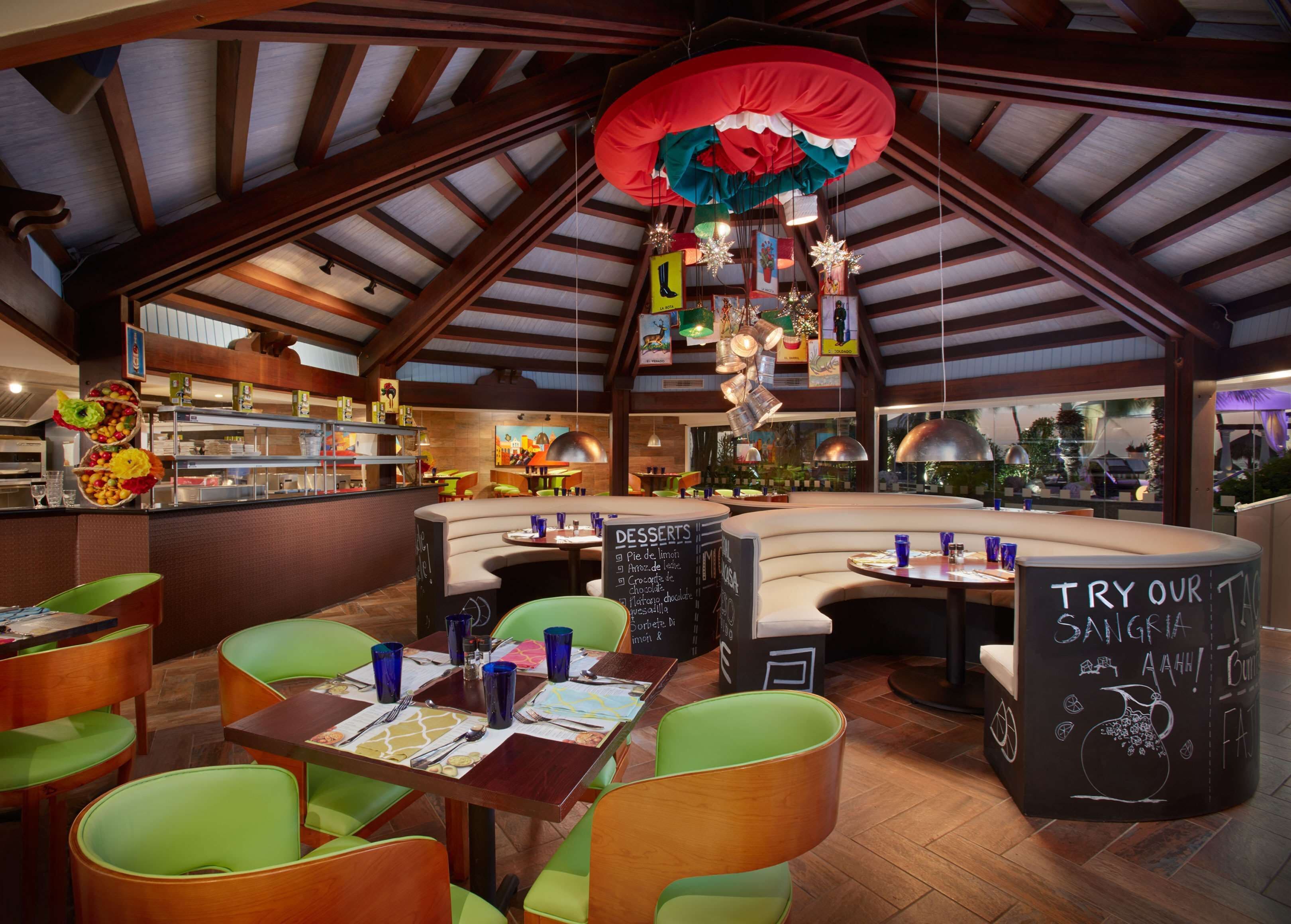 A restaurant with a high ceiling, wooden beams, and a variety of seating options including booths and tables. There is a chalkboard menu and a sign encouraging patrons to try the sangria.