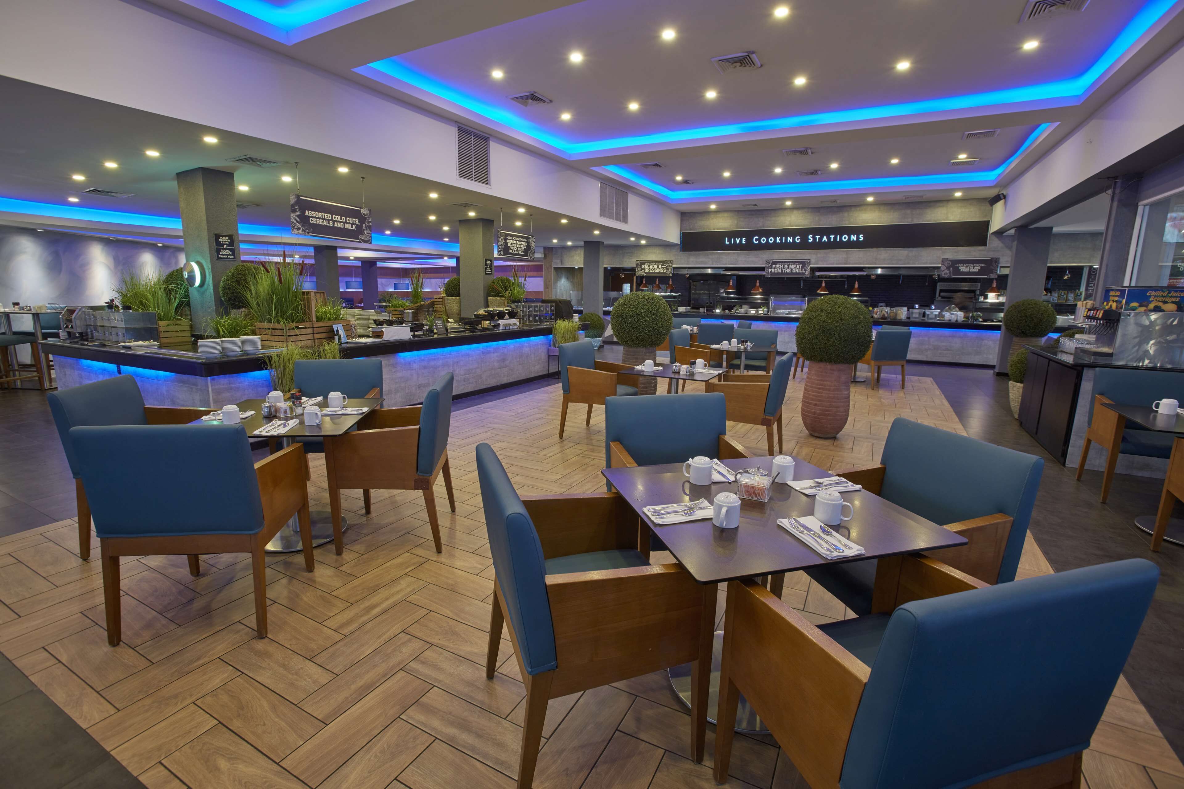 A modern restaurant with blue chairs, wooden tables, and a live cooking station.