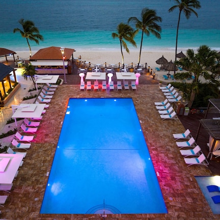 A pool area with lounge chairs and umbrellas, surrounded by palm trees and a beach view.