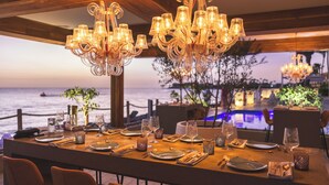 A dining table set for a meal with a view of the sea, elegant chandeliers, and a wooden ceiling.