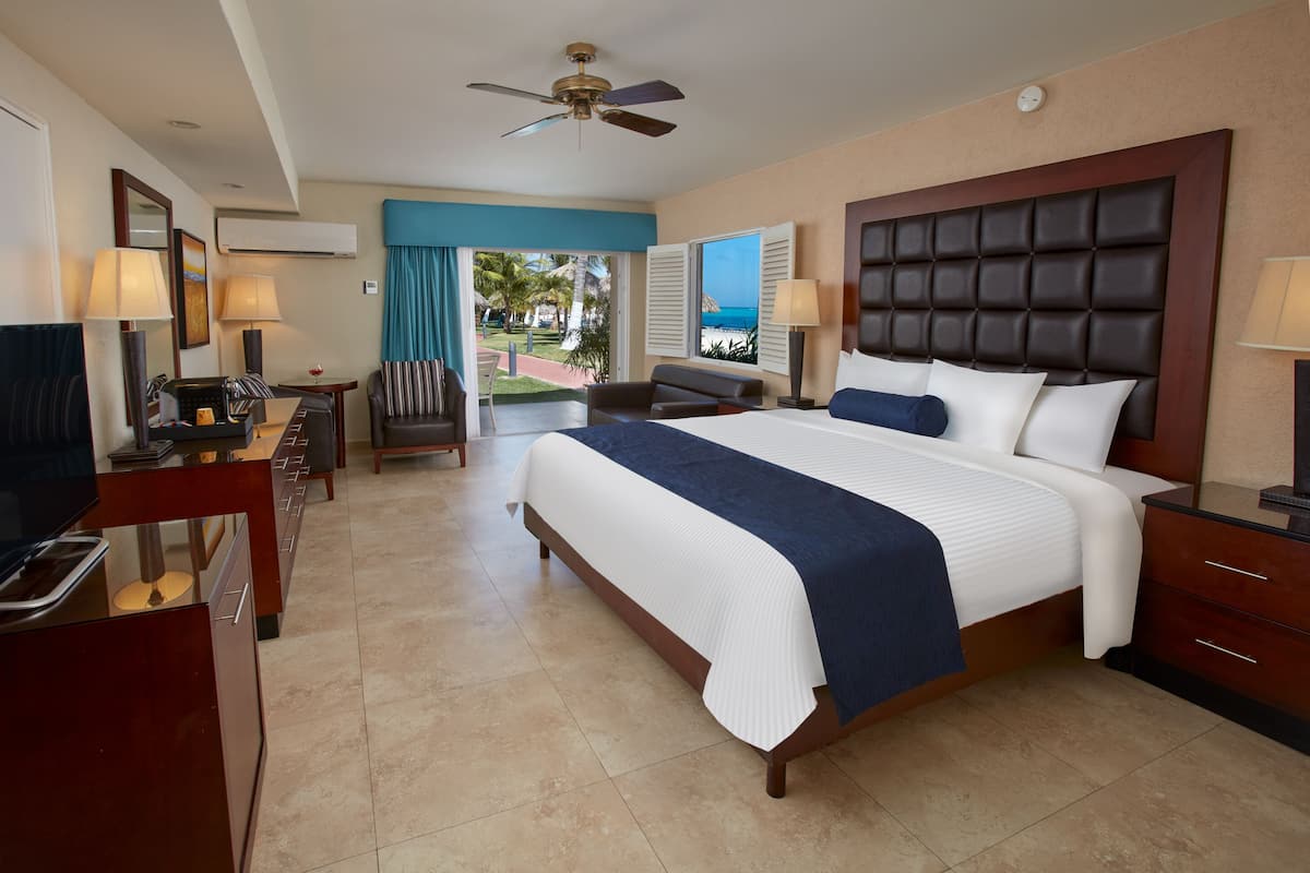 A hotel room with a large bed, a desk with a TV, a chair, and a view of the beach.