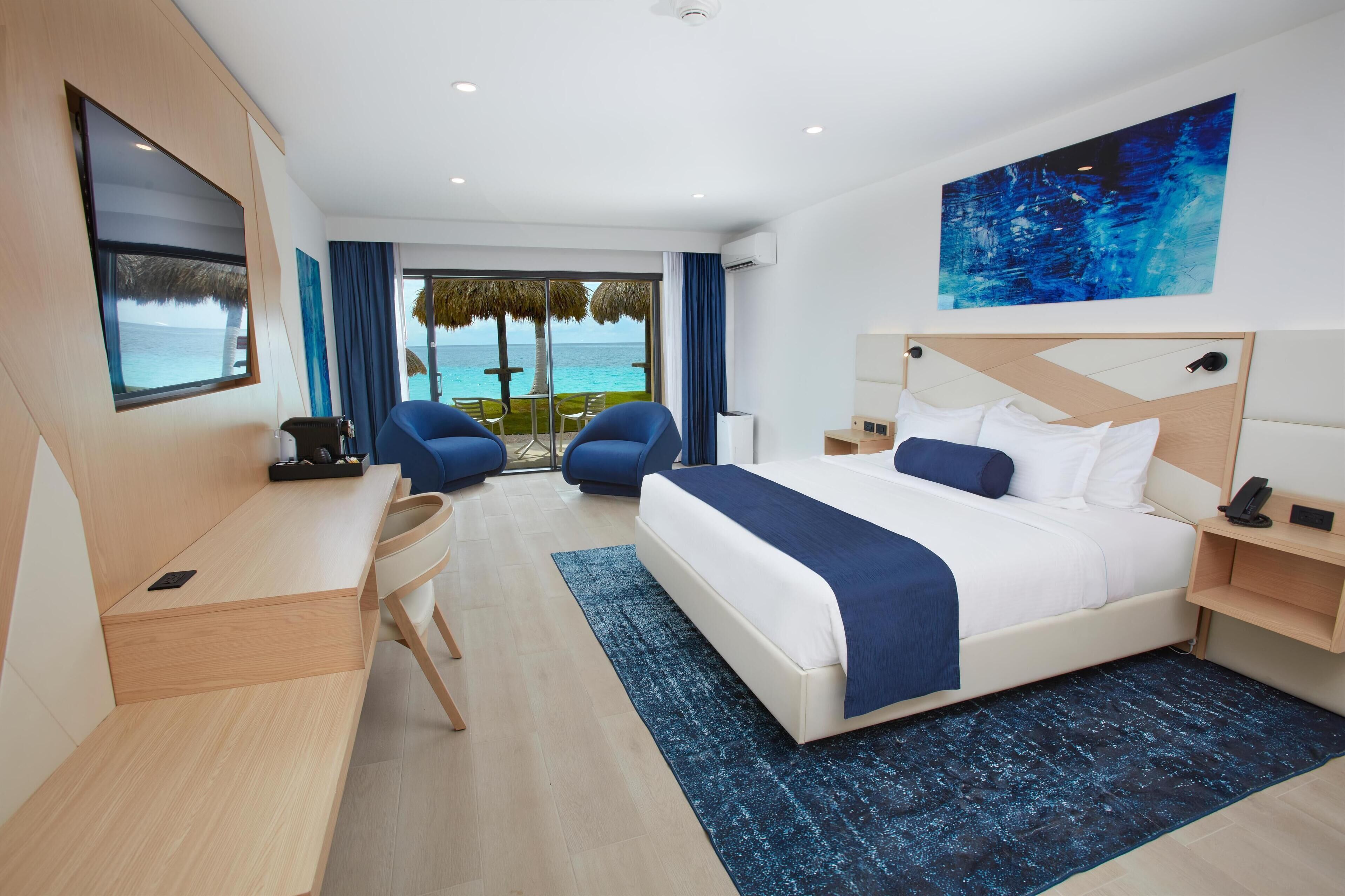 A modern hotel room with a large bed, a desk, and a chair. There is a view of the ocean through the window.