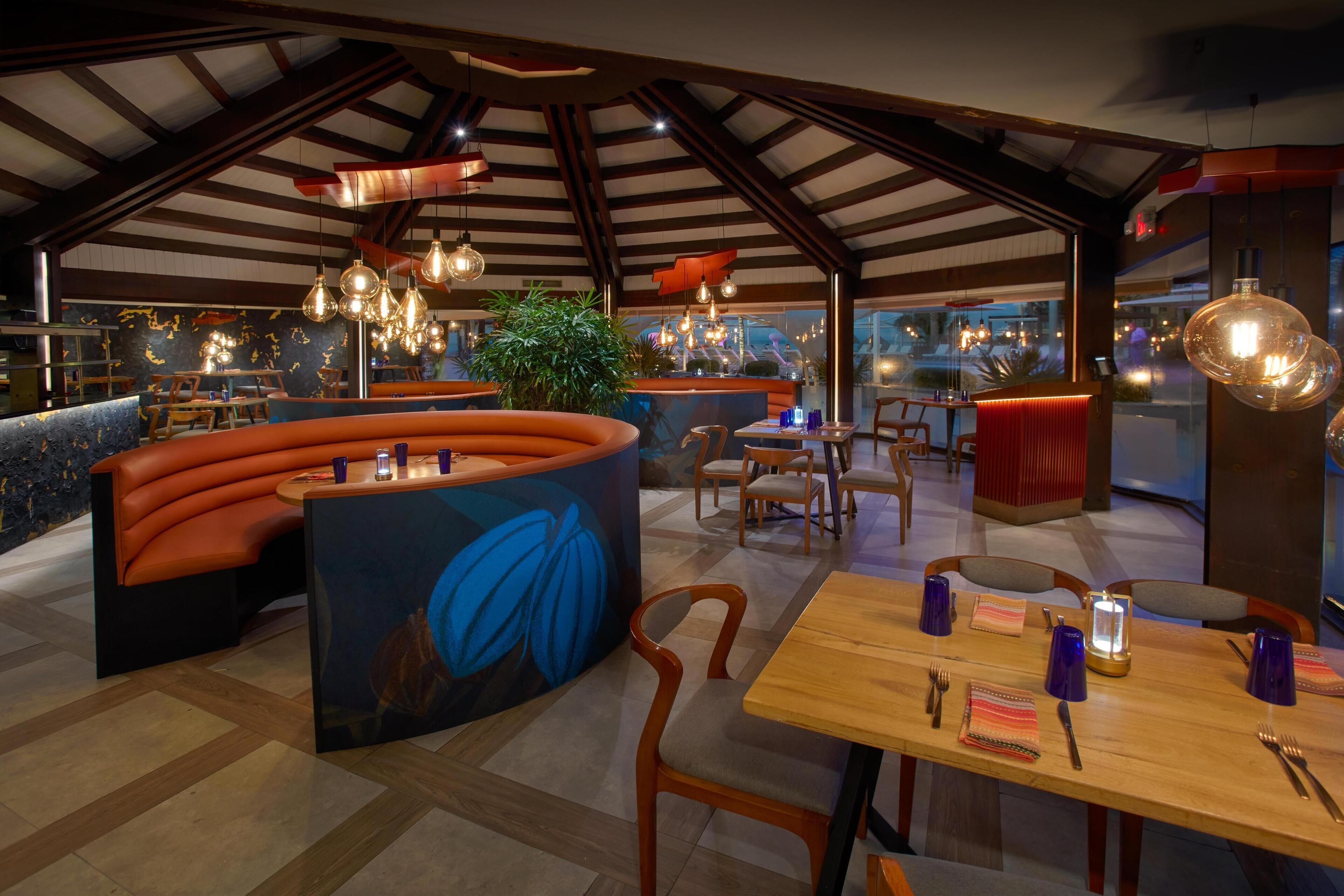 A restaurant with a high ceiling, exposed beams, and modern lighting fixtures. There are seating areas with orange booths and wooden tables.