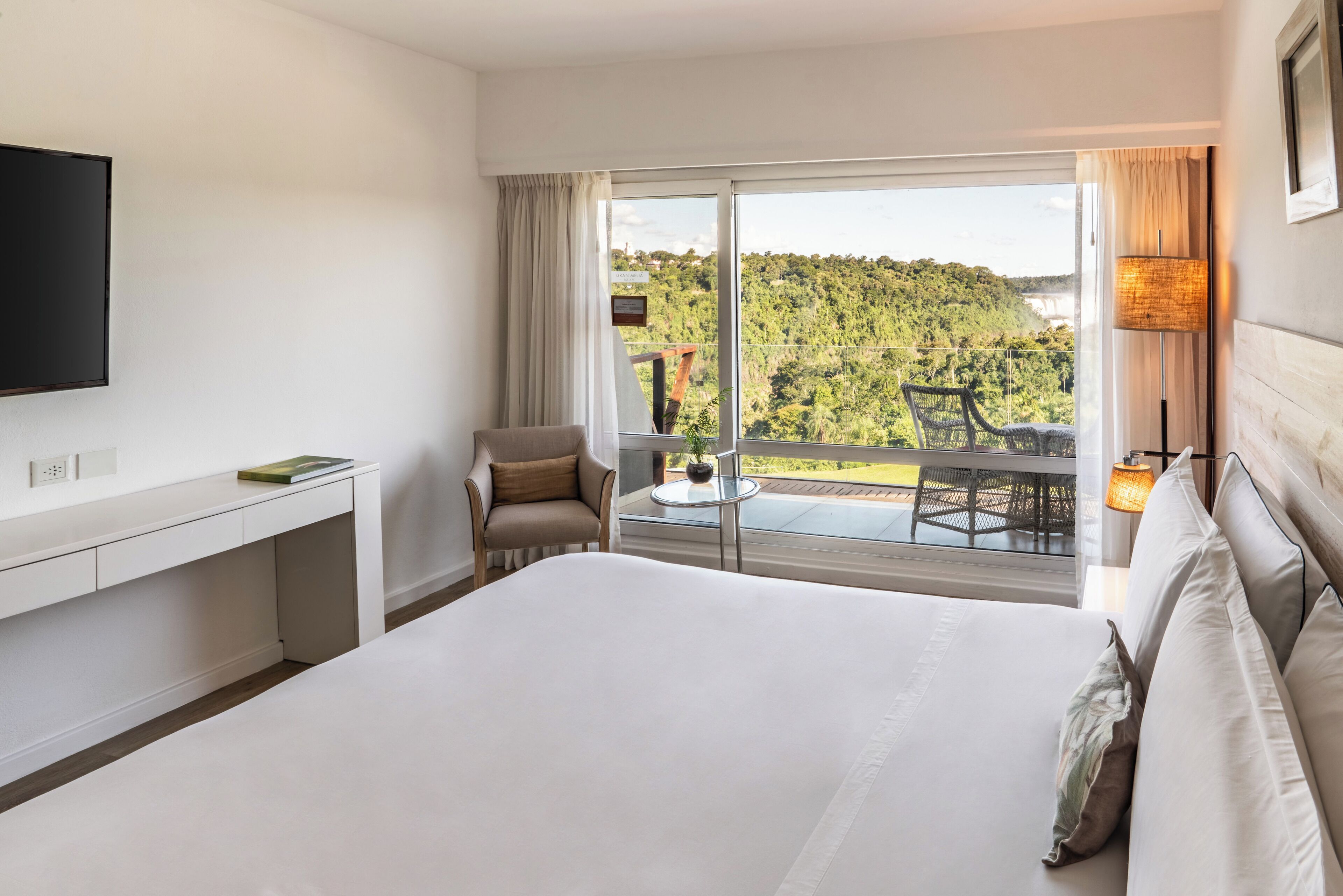 A hotel room with a large bed, a TV, a desk, a chair, and a balcony with a view of a forested area.