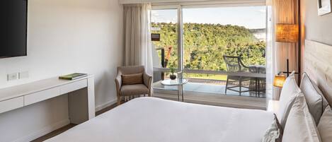 A hotel room with a large bed, a TV, a desk, a chair, and a balcony with a view of a forested area.