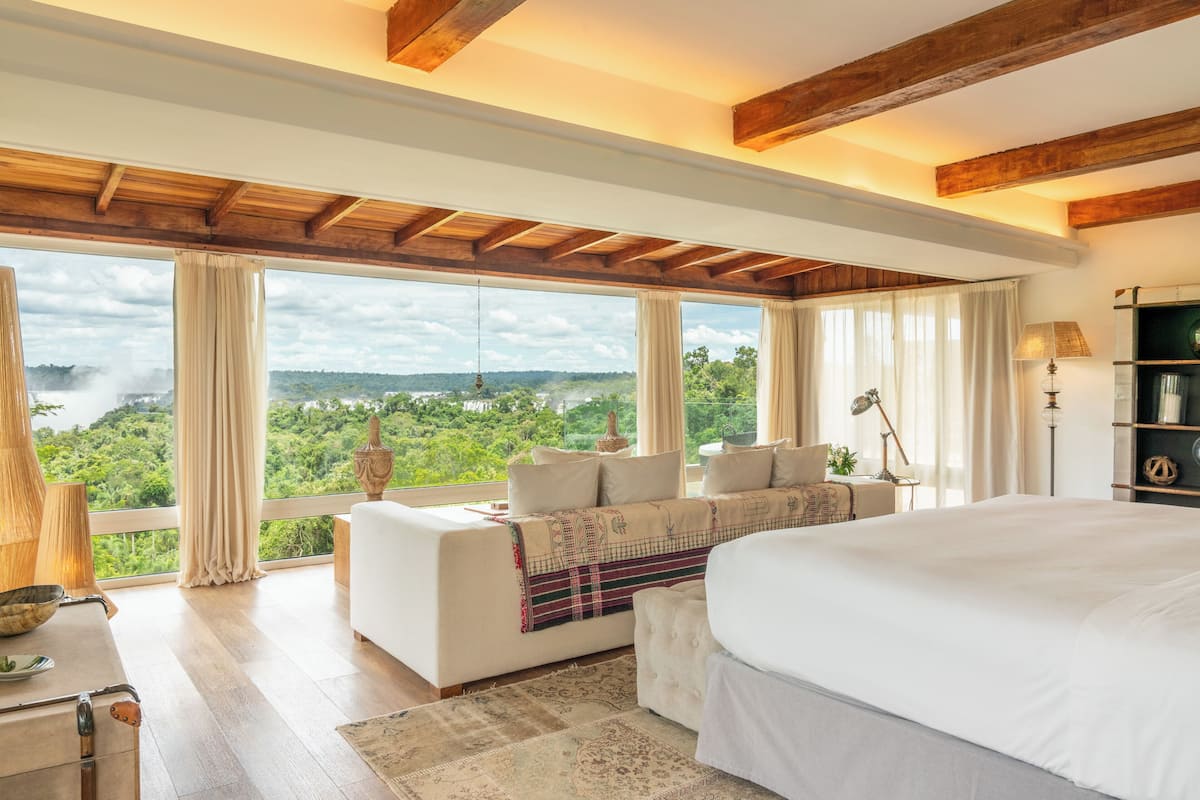 A spacious bedroom with a large bed, a sofa, and a view of a lush landscape through large windows.