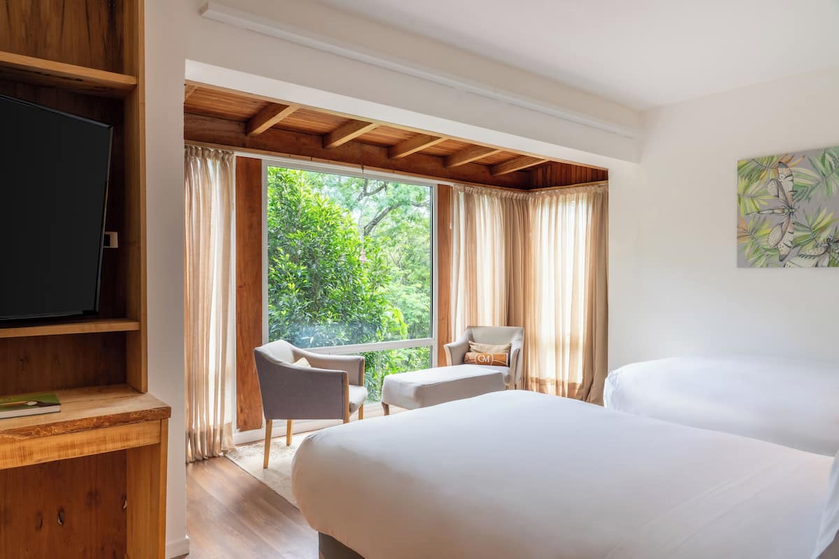 A bedroom with two beds, a TV, a desk, and a chair. There is a window with curtains and a view of greenery outside.