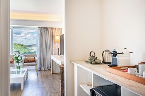 A modern hotel room with a dining area, a kitchen with a coffee maker, and a view of greenery.