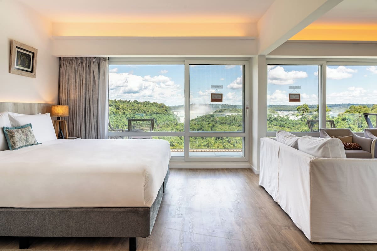 A hotel room with a large bed, a sofa, and a view of a forested area through large windows.