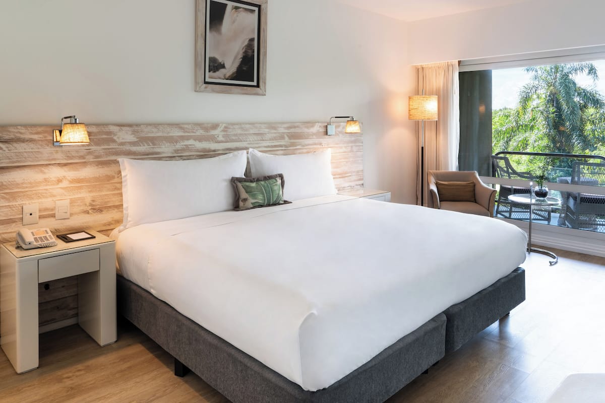 A hotel room with a large bed, two bedside lamps, a nightstand with a phone, a window with a view of greenery, and a balcony with a chair and table.