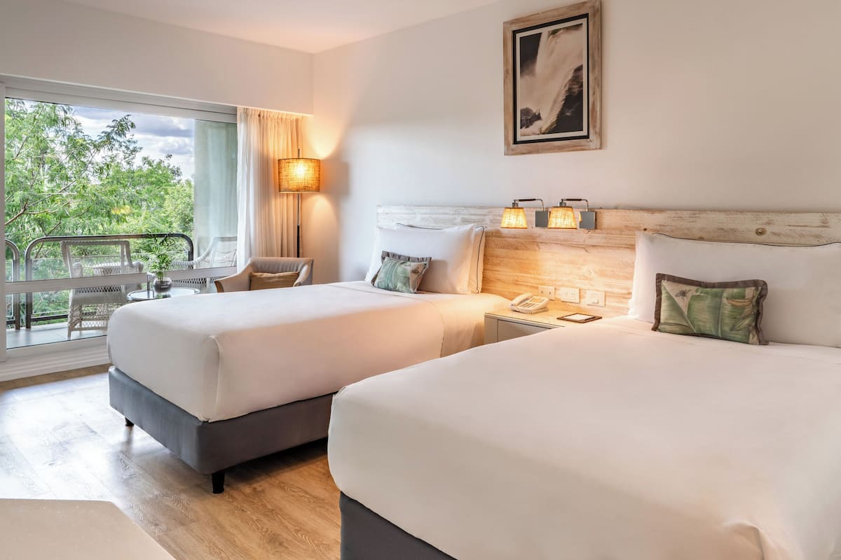 A hotel room with two beds, a balcony with a table and chairs, and a large window with curtains.