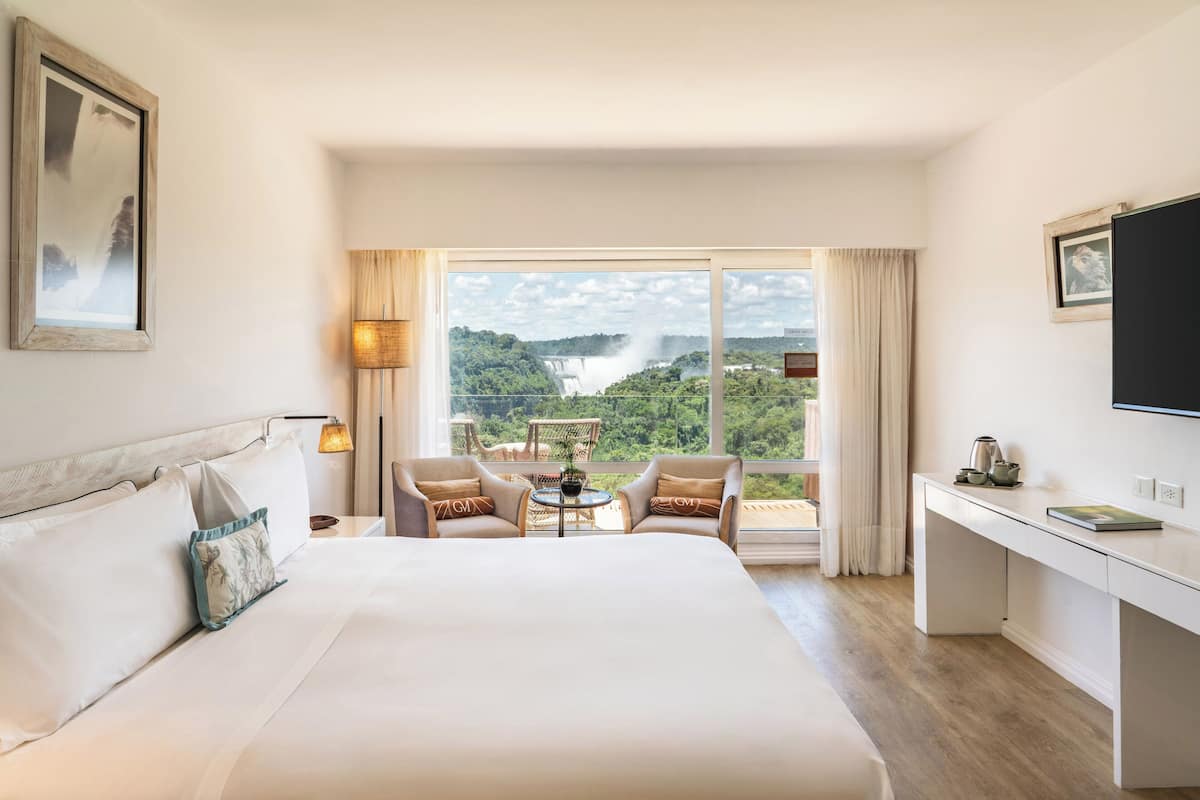 A hotel room with a large bed, two armchairs, a desk, and a view of a waterfall.