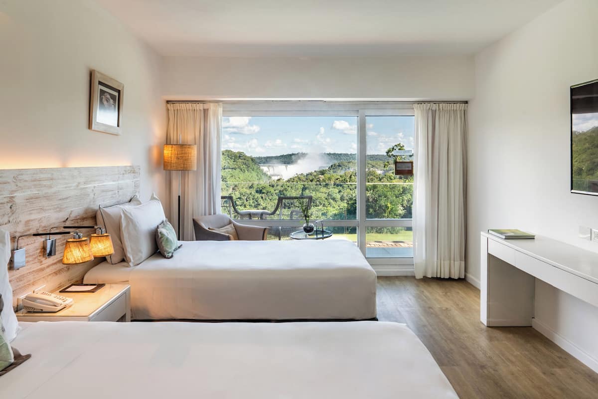 A hotel room with two beds, a desk, a chair, and a large window offering a scenic view of a waterfall and greenery.
