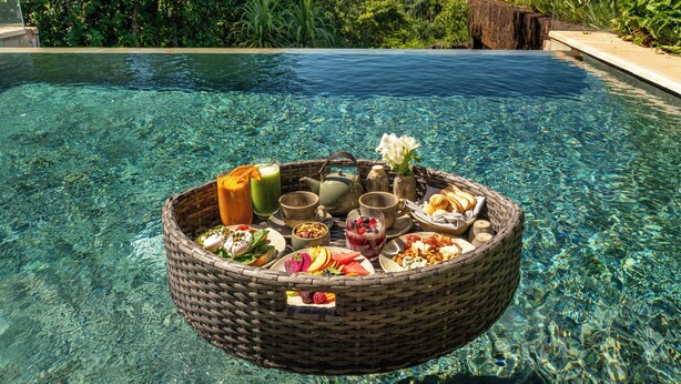 Ontbijt op een drijvende plank met een mand vol eten en drinken, een theepot en een bloem, bij een helder zwembad met uitzicht op een waterval.