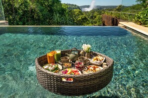 A floating breakfast setup with a basket containing food and drinks, a teapot, and a flower, by a clear pool with a waterfall view. - Gran Melia Iguazú (Puerto Iguazú)
