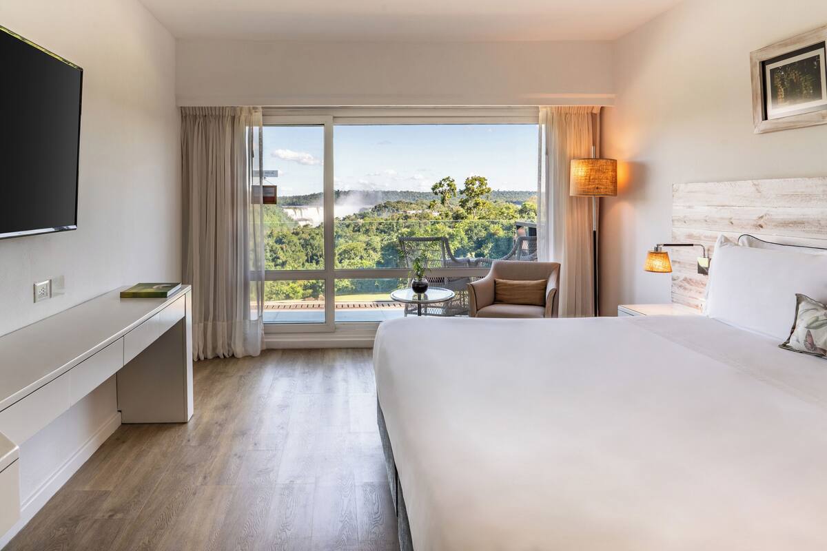 A hotel room with a large bed, a desk, a chair, a TV, a lamp, and a view of a lush landscape through the window.