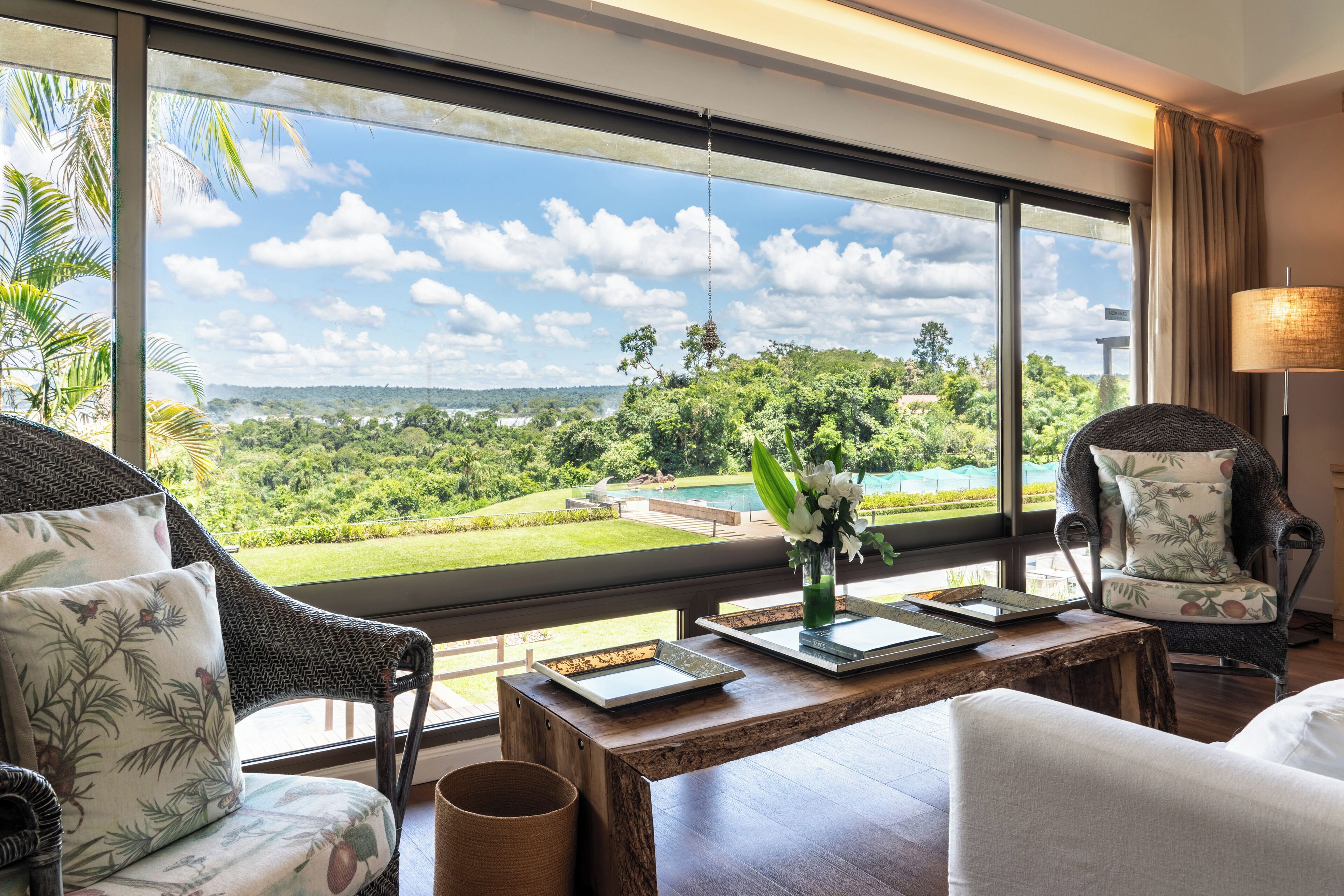 Una habitación con un ventanal que da a un paisaje exuberante, una mesa de madera con un florero y dos sillas de mimbre.