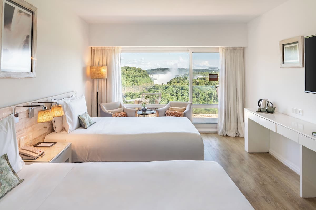 A hotel room with two beds, a desk, a chair, and a view of a waterfall.