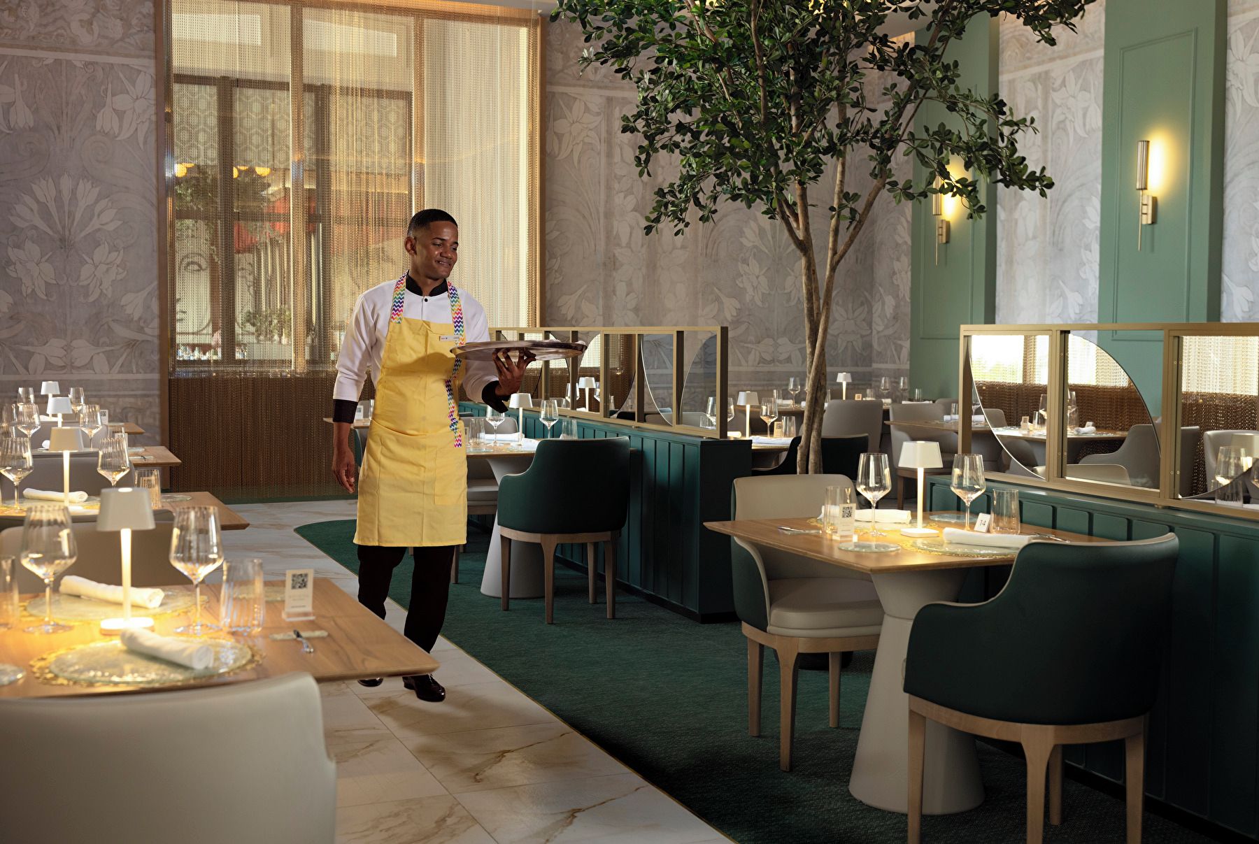 A restaurant interior with tables set for dining, a waiter in uniform, and a large mirror reflecting the room.