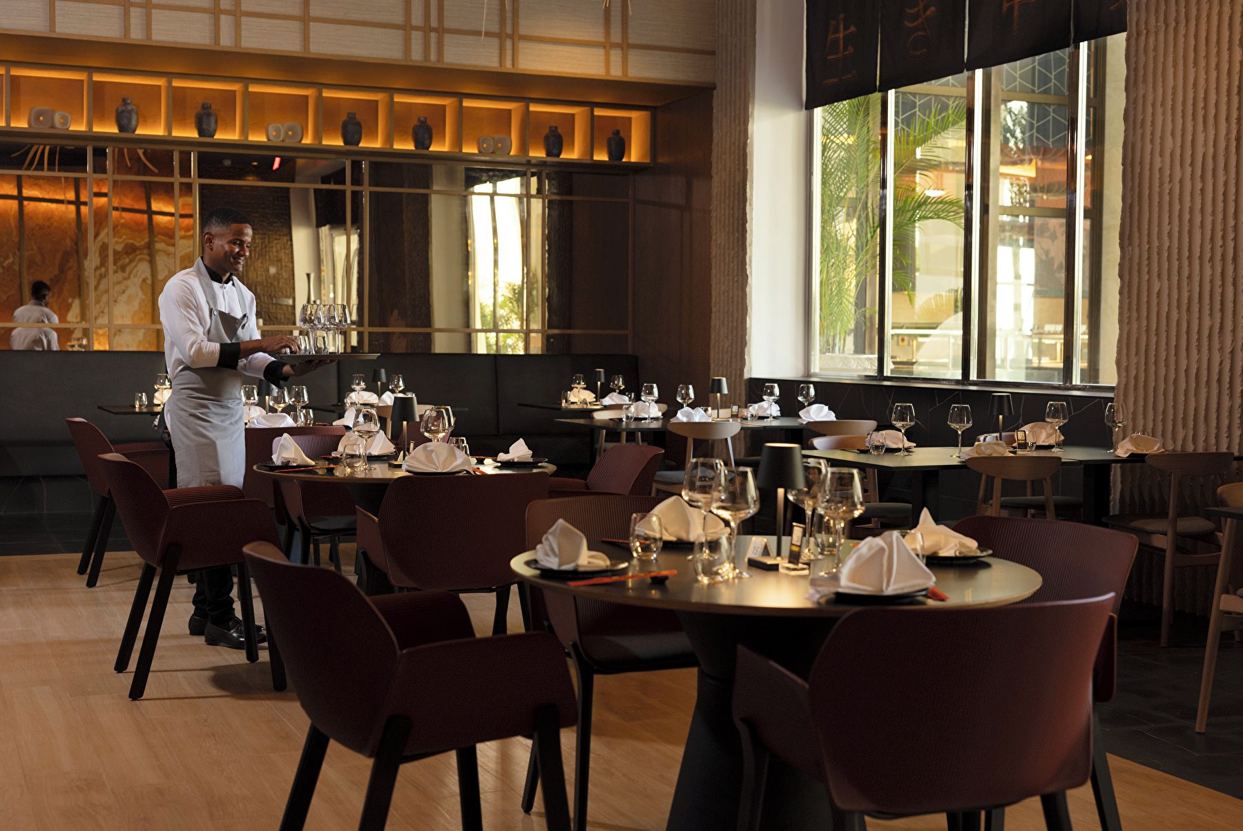 A restaurant interior with tables set for dining, a waiter attending to a table, and a view of the outdoors through large windows.