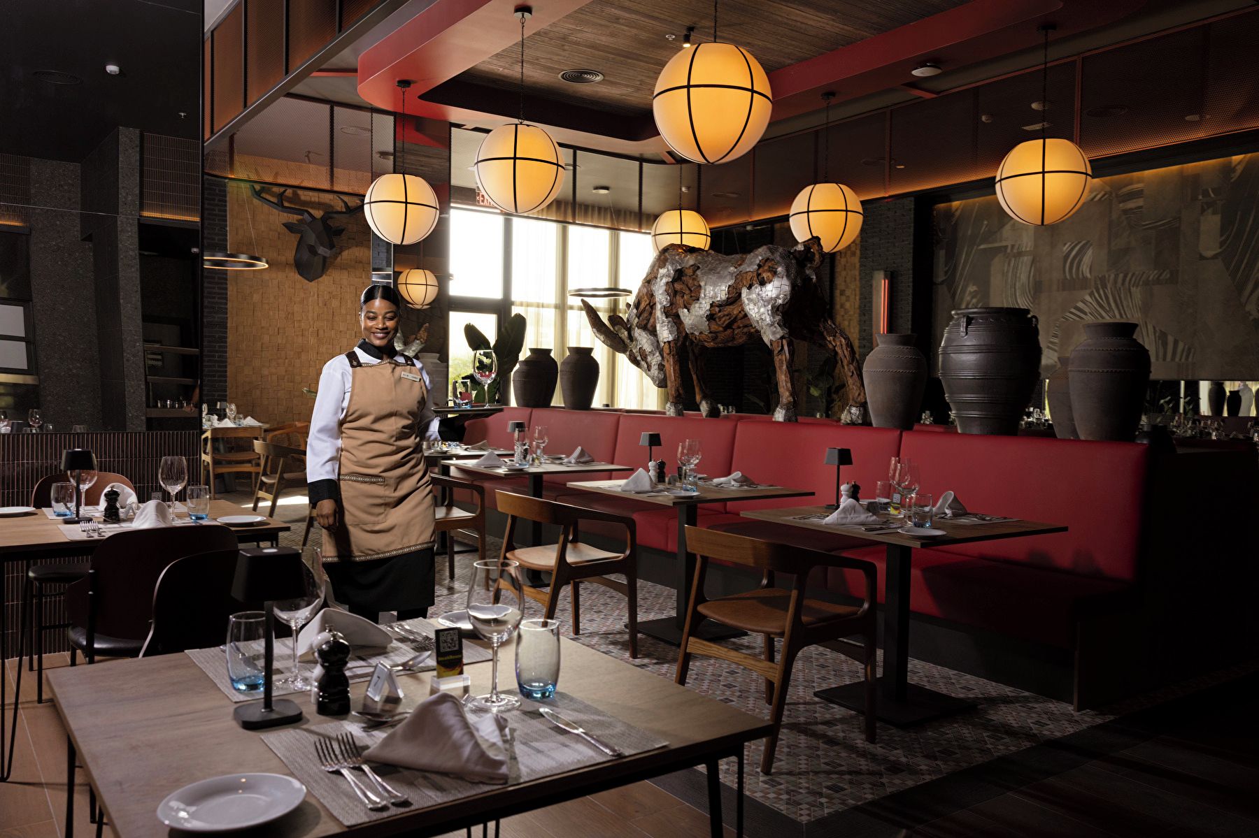 A restaurant interior with tables set for dining, a waiter, and decorative elements like a bull sculpture and hanging lamps.
