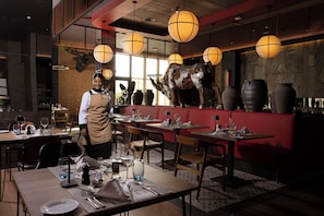 A restaurant interior with tables set for dining, a waiter, and decorative elements like a bull sculpture and hanging lamps. - Hotel Riu Palace Aruba - All Inclusive (Noord)