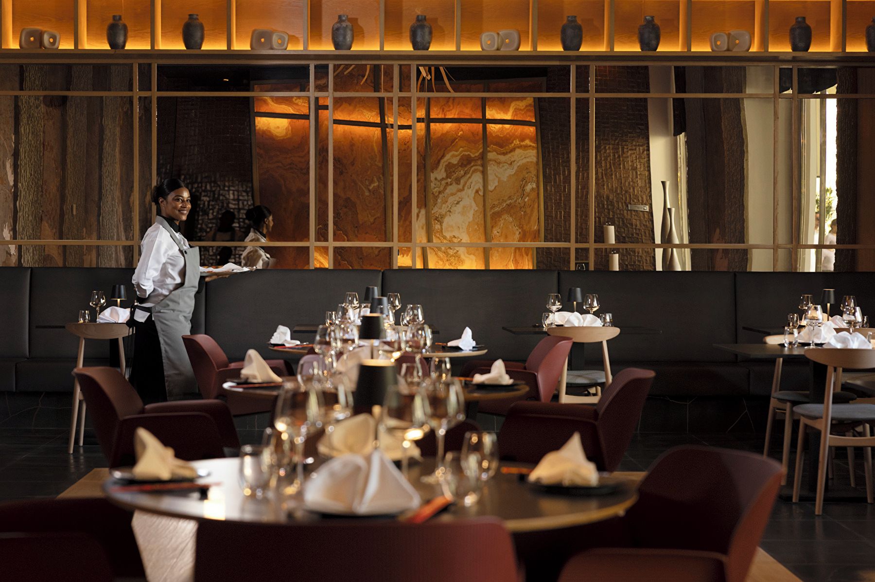 L’intérieur d’un restaurant, avec des tables dressées pour le service, un serveur en uniforme et un éclairage d’ambiance chaleureux.