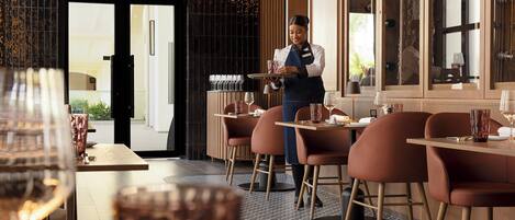Un interior de restaurante con un camarero atendiendo, mesas con sillas y vistas a la ciudad a través de las ventanas.