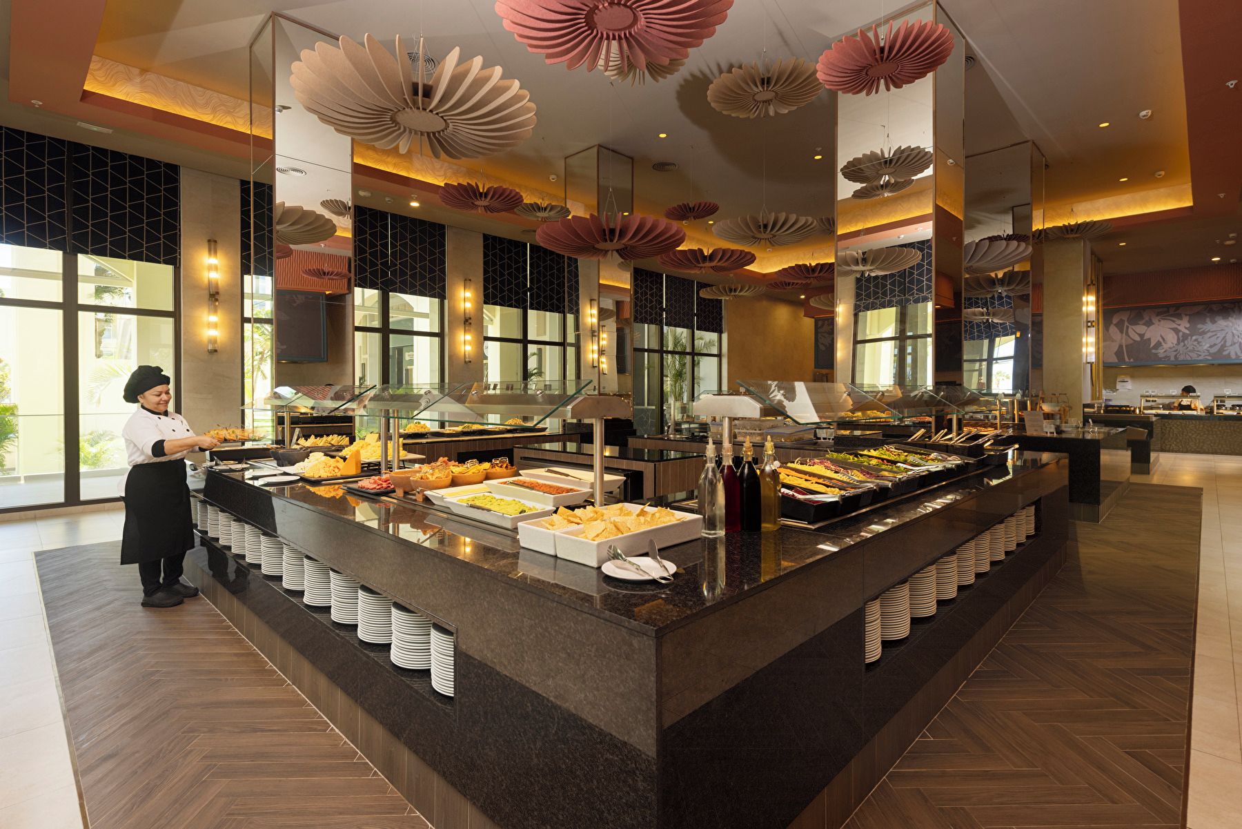 A buffet area with a variety of food options, a buffet server, and a modern interior design with large windows and decorative ceiling elements.