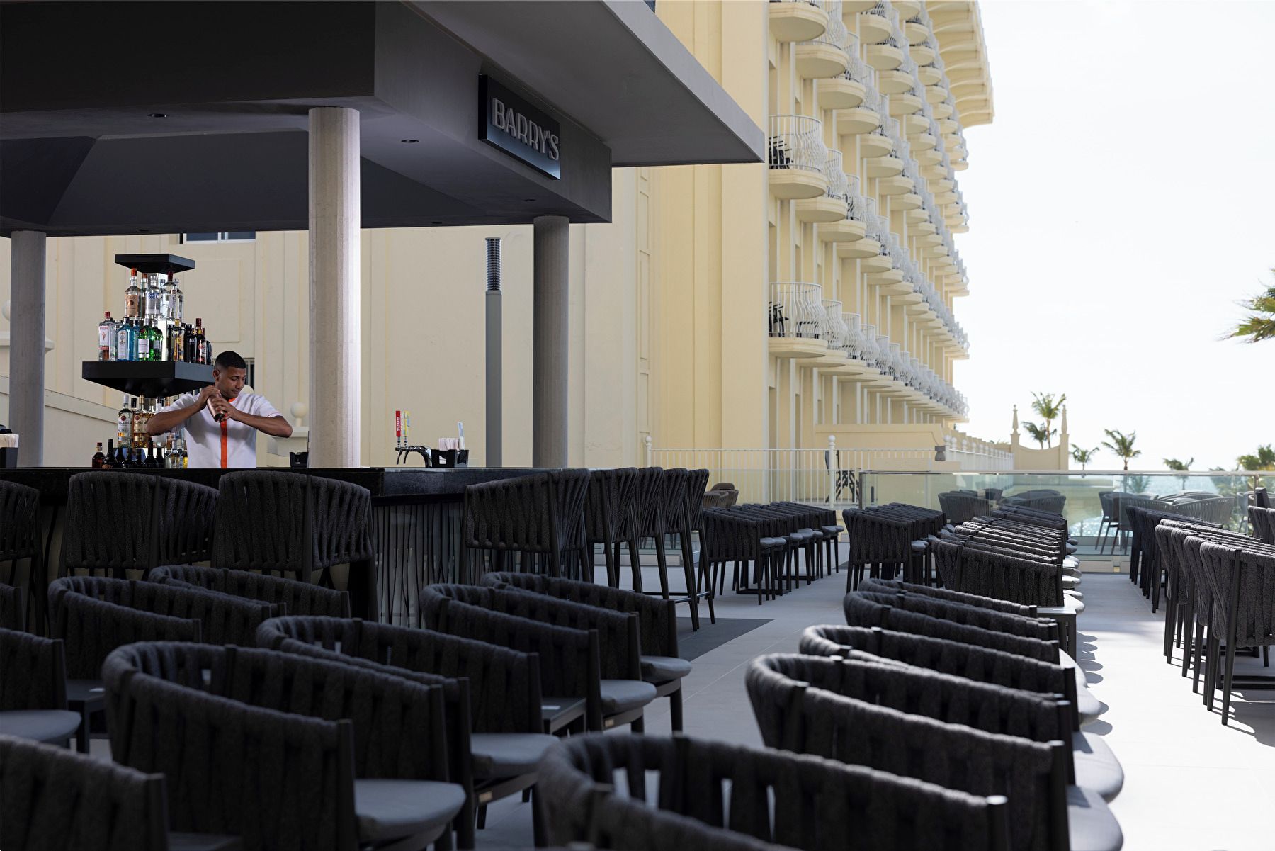 A bar area with a bartender, a liquor display, and outdoor seating with tables and chairs.