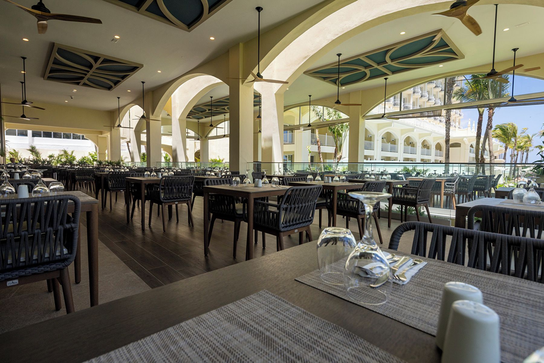 Un restaurant doté de nombreuses tables et chaises, de grandes fenêtres et offrant une vue sur un immeuble avec des balcons.