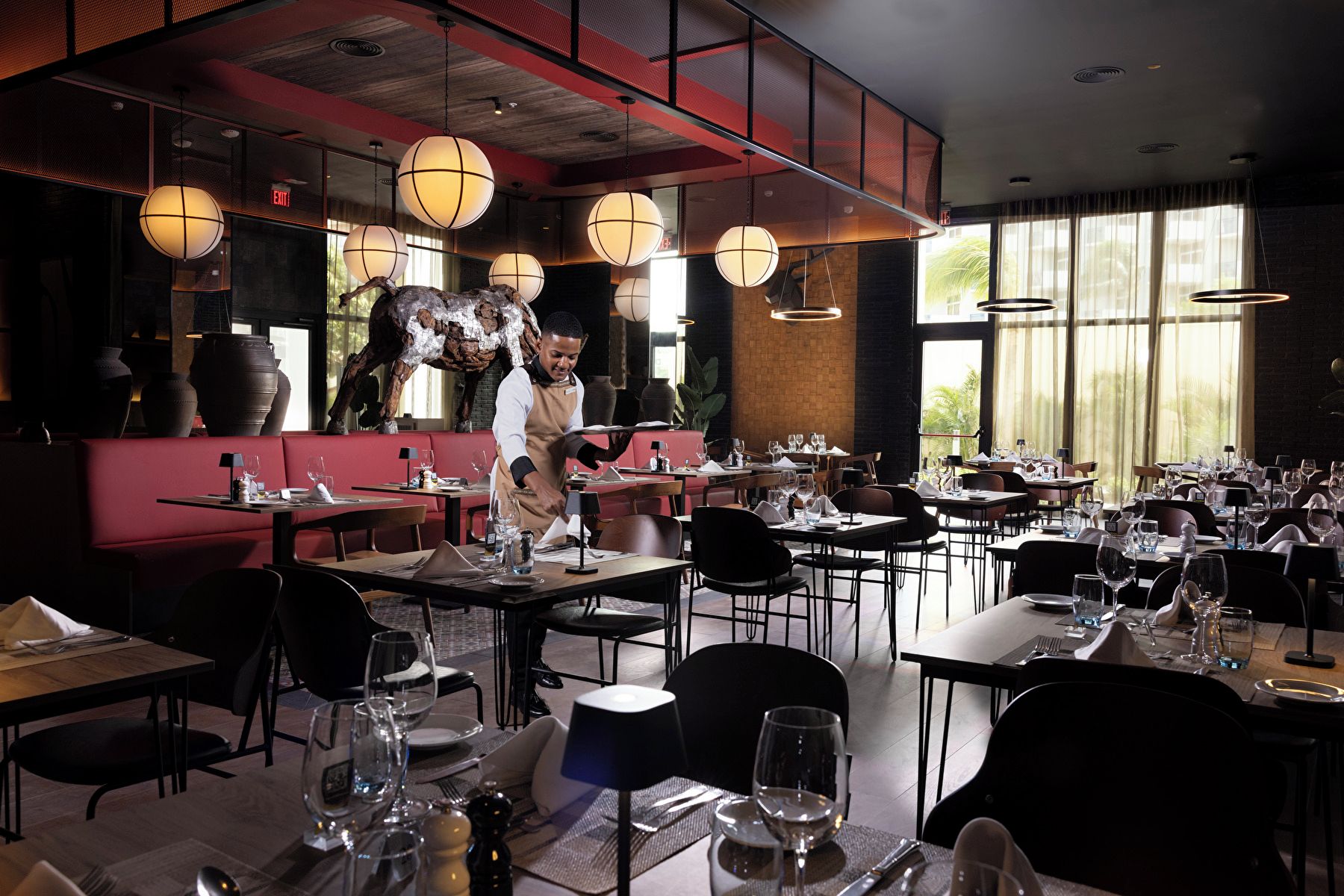 A restaurant interior with tables set for dining, a waiter in service, and a large decorative sculpture.