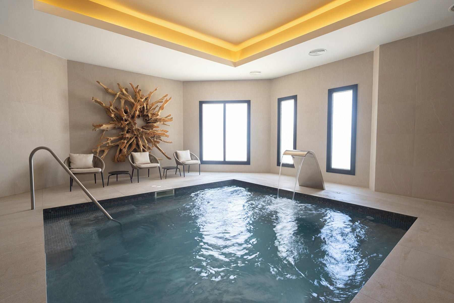 A modern indoor swimming pool with a clear water surface, surrounded by lounge chairs and a large wooden sculpture.