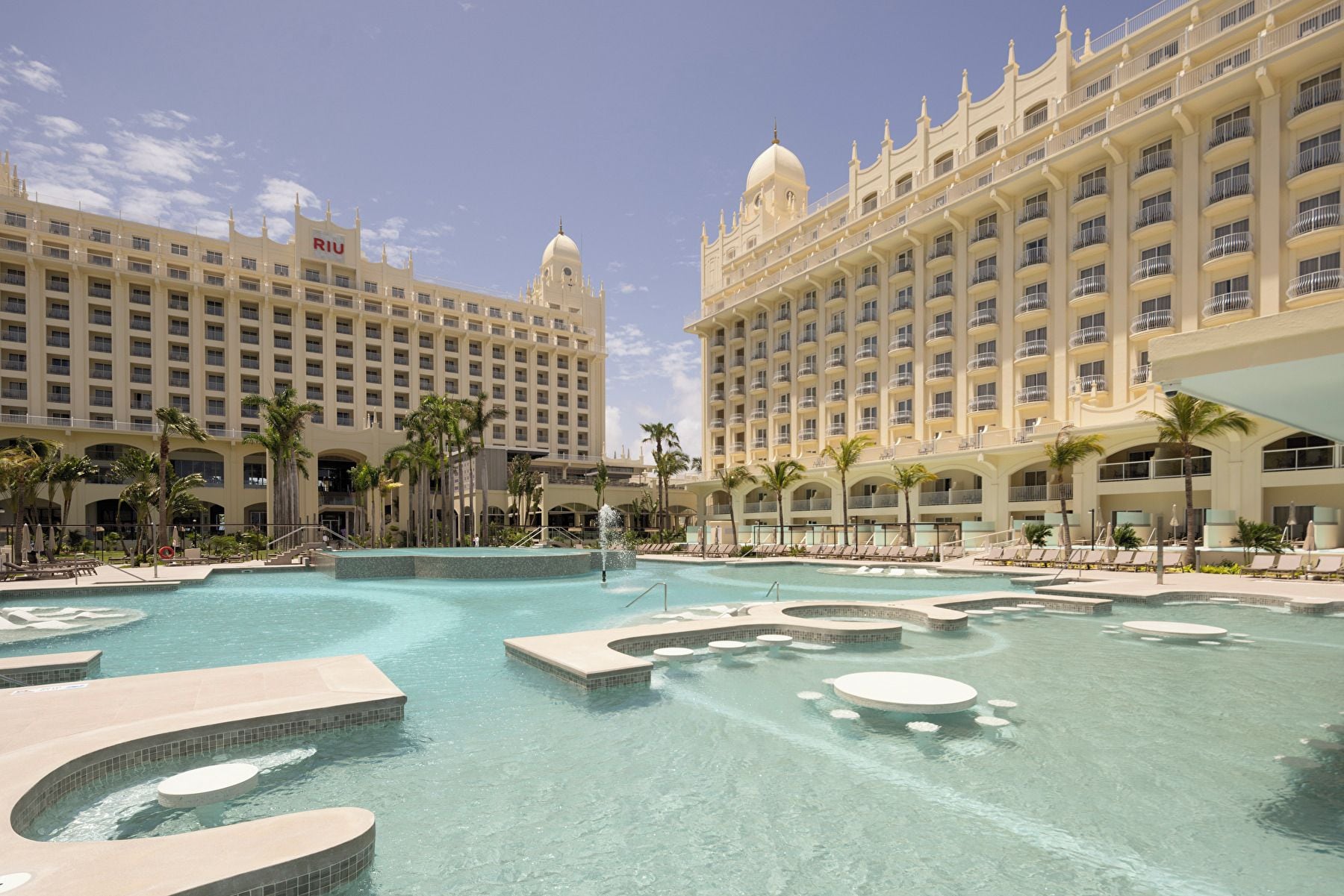 A hotel with a large central pool, multiple balconies, and palm trees.
