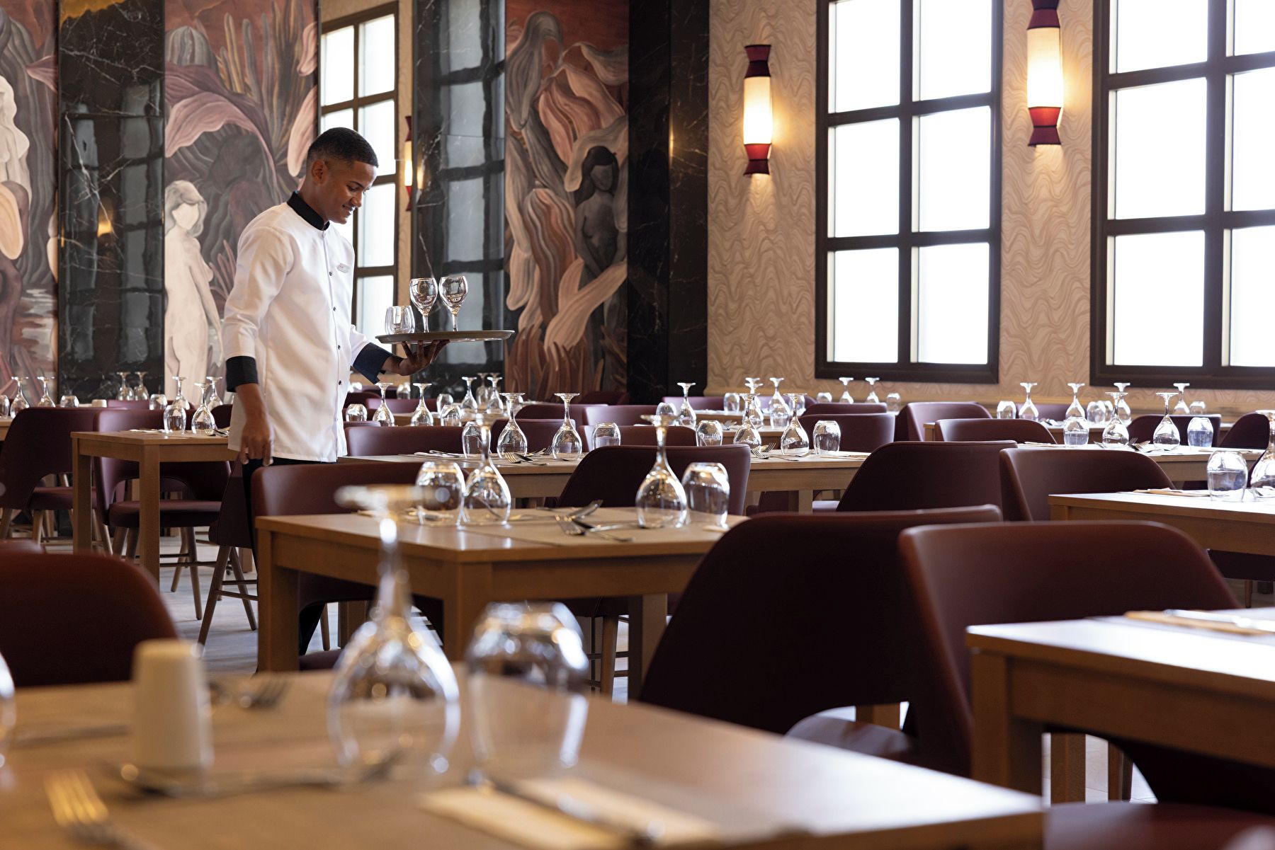 A restaurant interior with tables set for dining, a waiter carrying a tray, and decorative wall art.