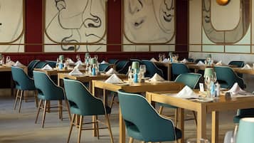 A restaurant with tables set for dining, teal chairs, and a wall with abstract art.