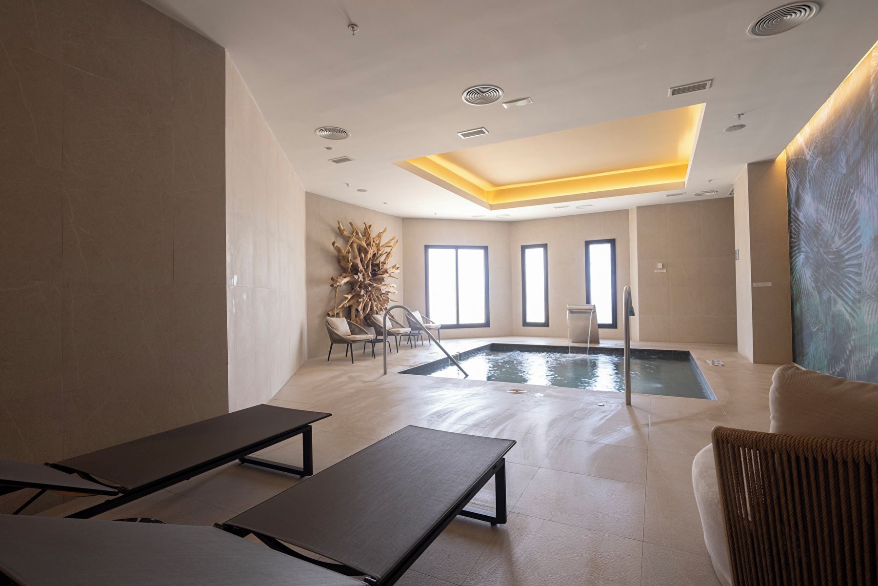 Un espace piscine intérieur moderne, doté de chaises longues, d’une grande sculpture en bois et offrant une vue sur la piscine.