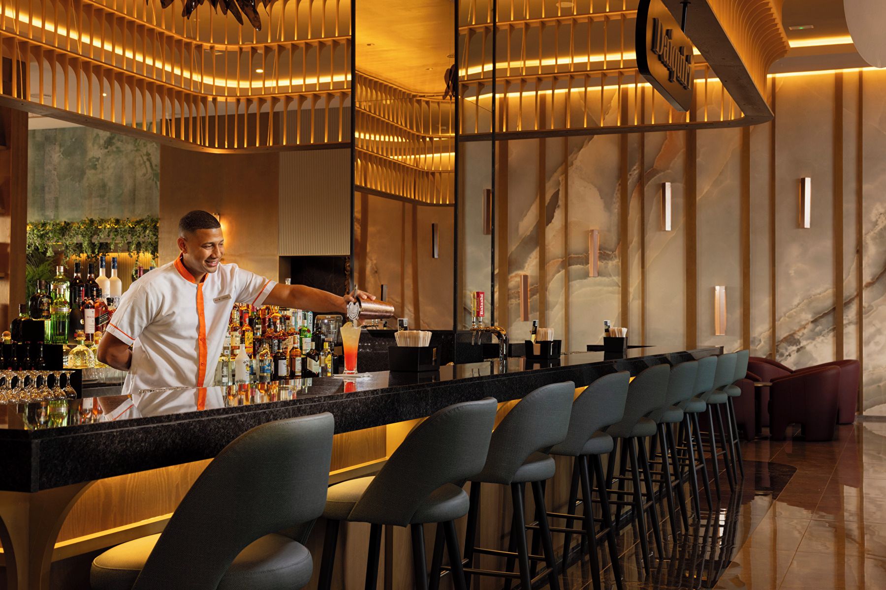 Zona de bar con un coctelero preparando una bebida, una barra larga con taburetes y un diseño interior moderno.